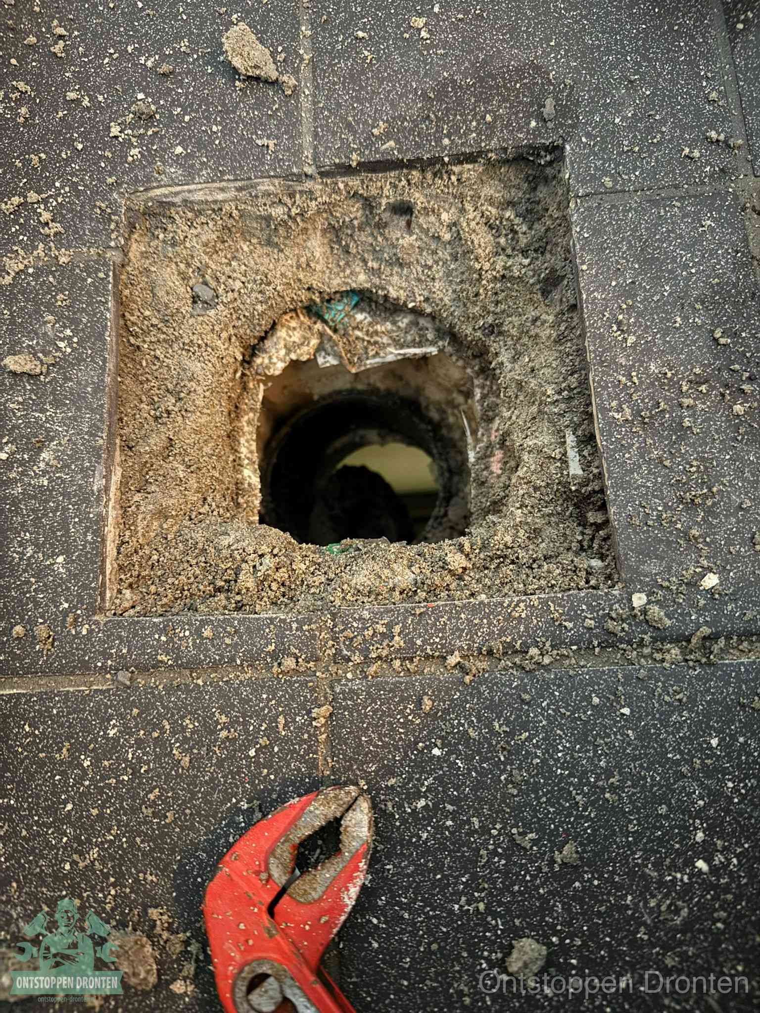 Een geopend rioolputje in een betontegelvloer met losgemaakt rooster en een zichtbare ronde afvoerbuis, het achterhalen waar de afvoerverstopping zit in Dronten