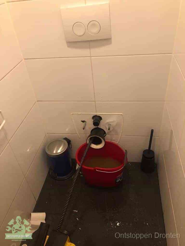 Een ontstopte wand-wc met verwijderde toiletpot waar een ontstoppingsveer in de wanduitgang verdwijnt, het achterhalen waar de verstopping zit in Dronten